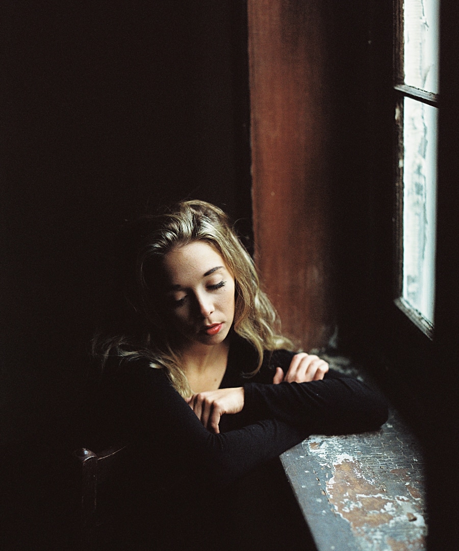 Senior girl sitting by a large window in a softly lit room, Morristown NJ senior portraits