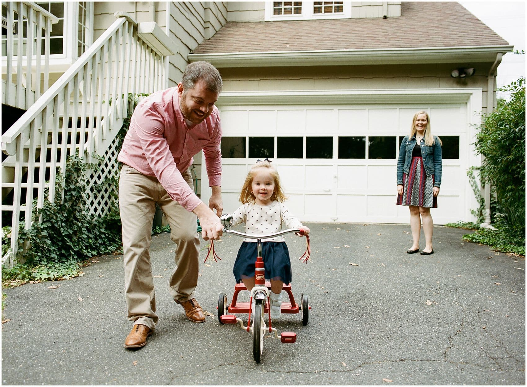 Outdoor lifestyle family portrait in Short Hills, NJ – little girl having fun riding a bike with her dad and mom looking on