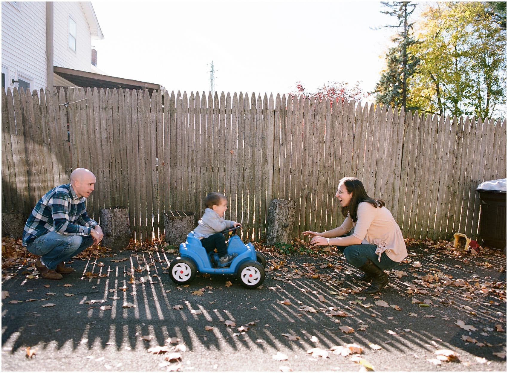 fun family photo of a little boy riding a blue car from mom to dad in front of the fence in NY
