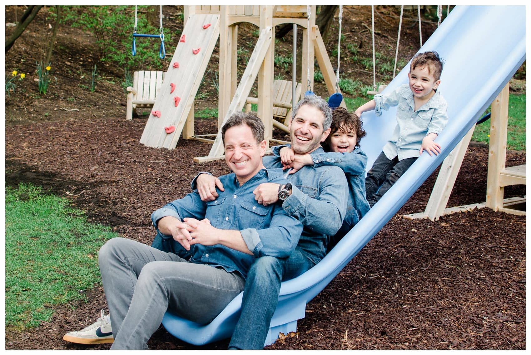 laughing fathers and sons are sliding down the blue slide at their family photos in Montclair NJ