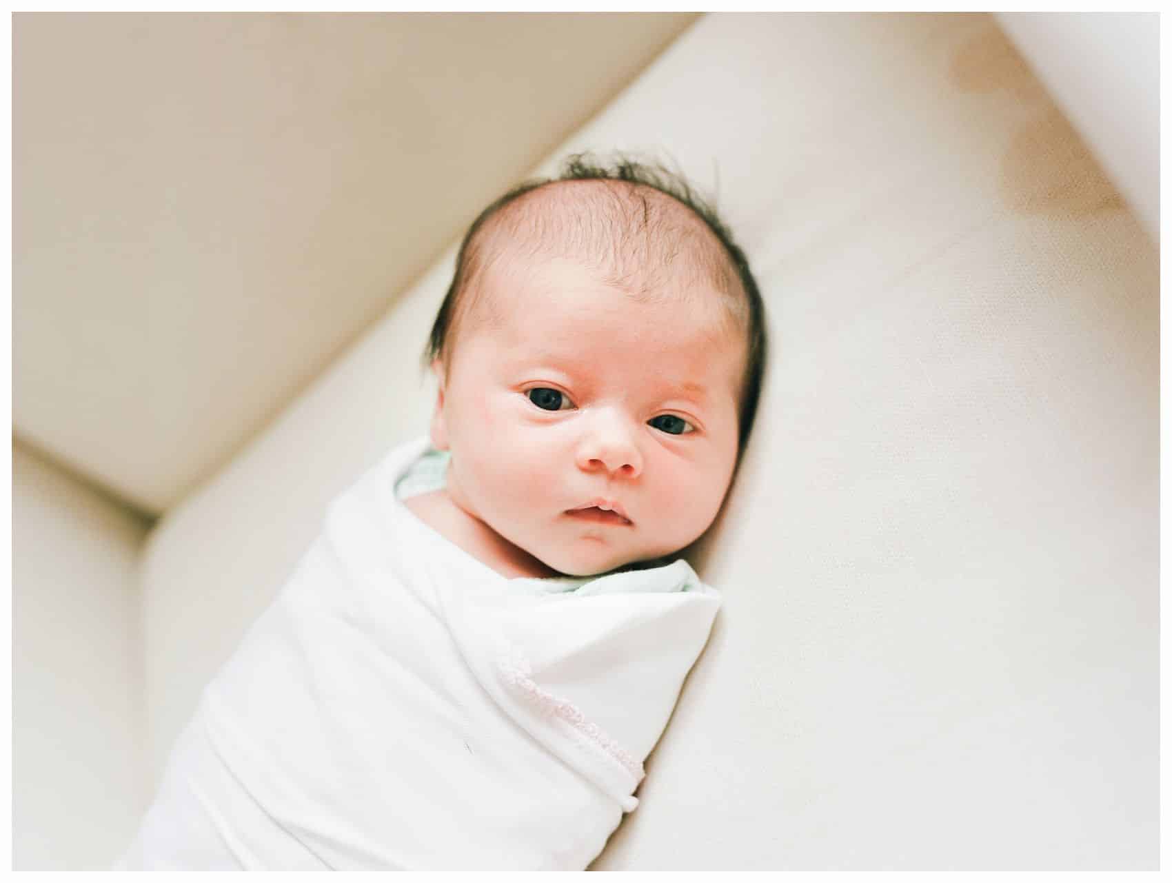 best newborn photographer taking a photo fo an awake newborn baby girl swaddled in a white blanket 