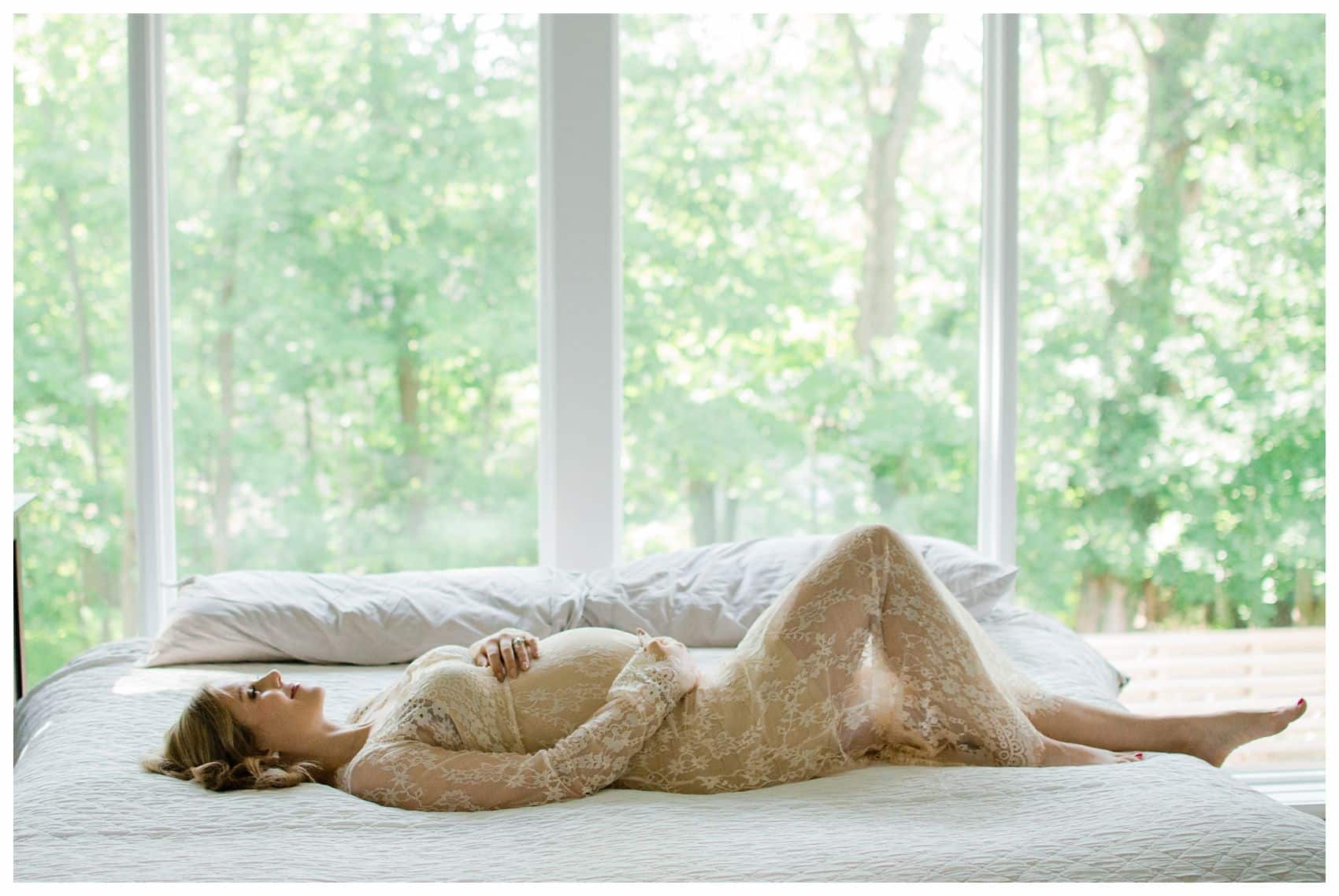 pregnant mom wearing lace dress laying blissfully on her white bed with trees in the background in Verona NJ