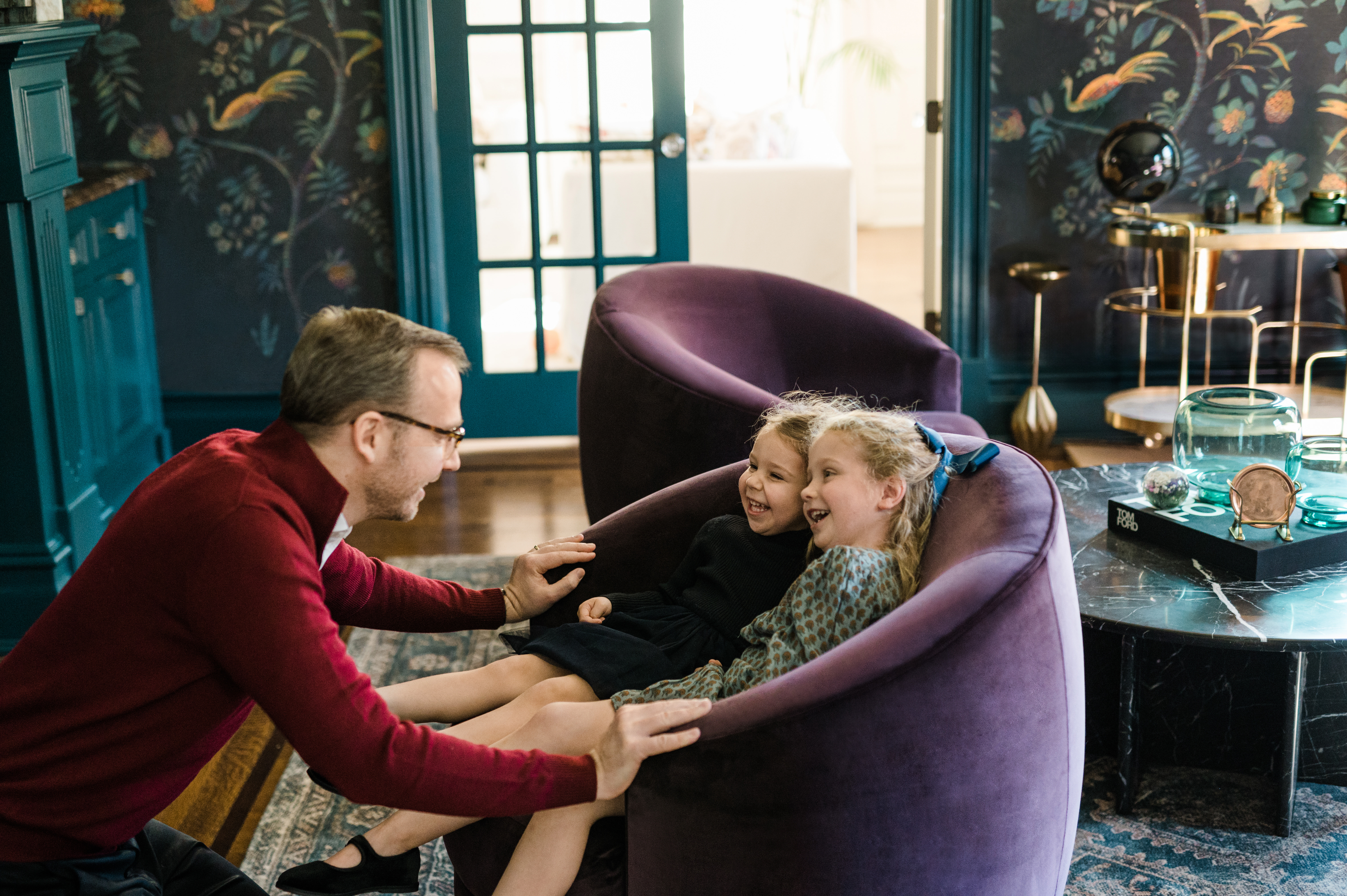 father is laughing with his two small daughters in their living room armchair